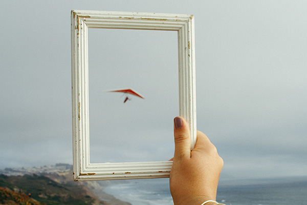 Eine Frauenhand hält einen weißen Bilderrahmen gegen den Himmel und rahmt damit einen roten Drachenflieger am Himmel ein